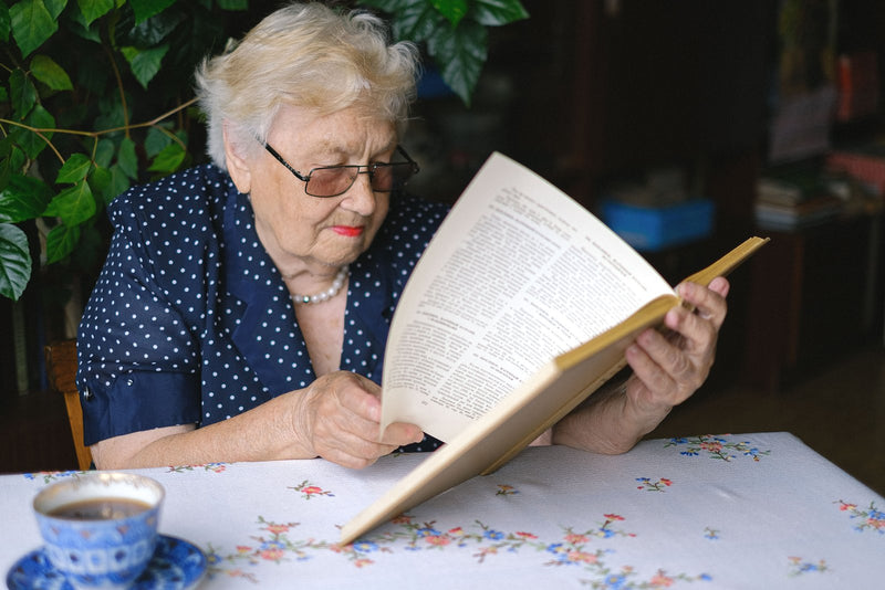 Older woman in glasses reading a book at her table with a cup of tea