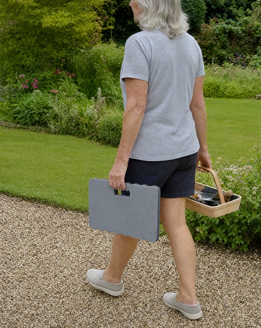 A woman carries the grey garden kneeler pad by its cut-out handle along a garden path