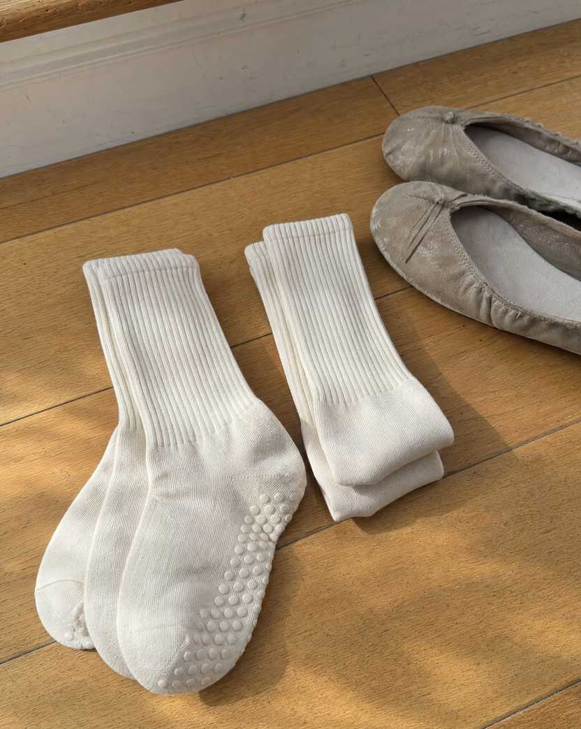 Two pairs of ivory grip socks folded on a wooden floor beside a pair of house shoes